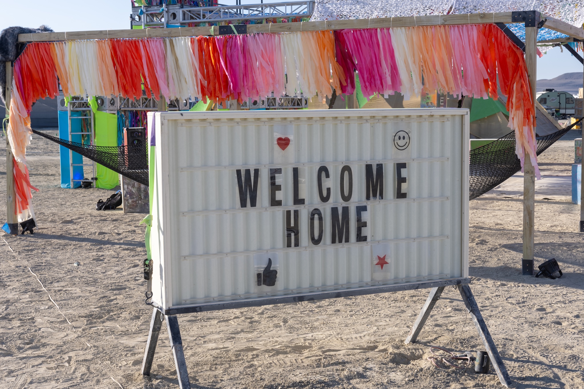 a sign in the desert spelling the words "welcome home" in front of a shade structure containing a hammock